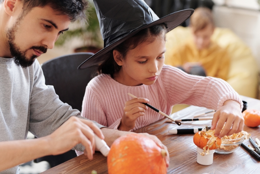 A young girl in a witches hat and her father decorating pumpkins