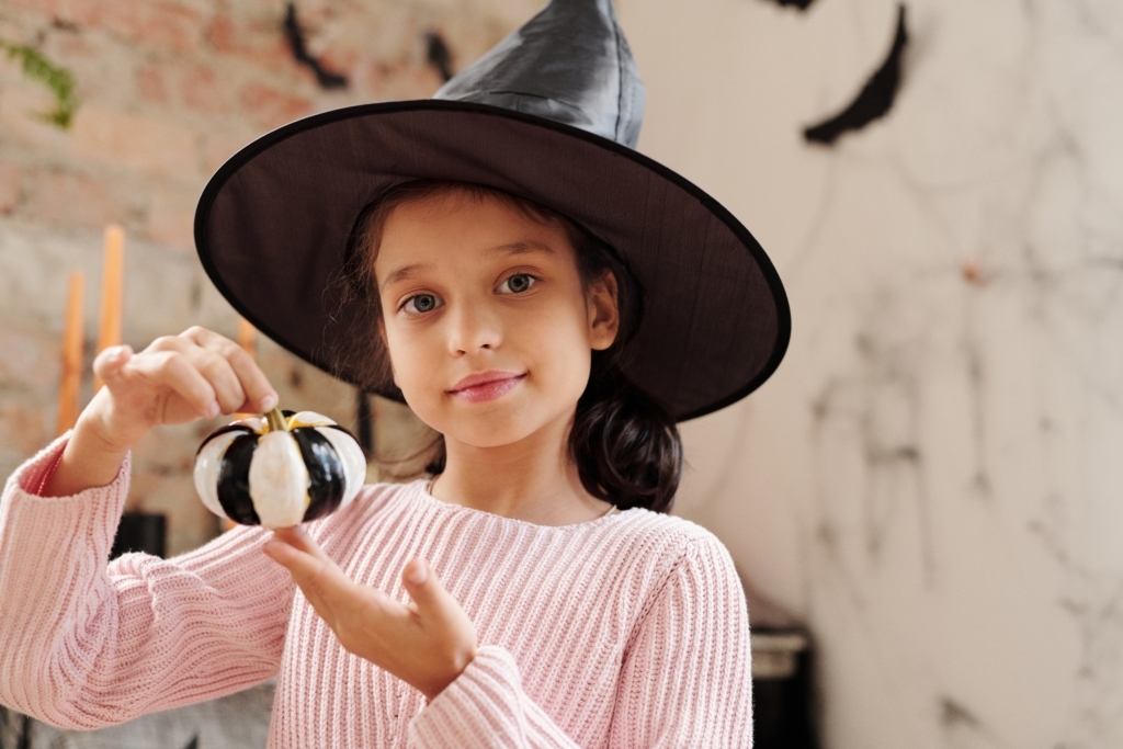 young girl in a witches hat holds a painted mini pumpkin