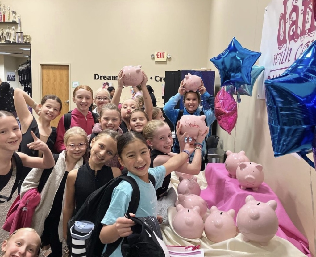 A group of young girls smile as they hold up and collect pink piggy banks.
