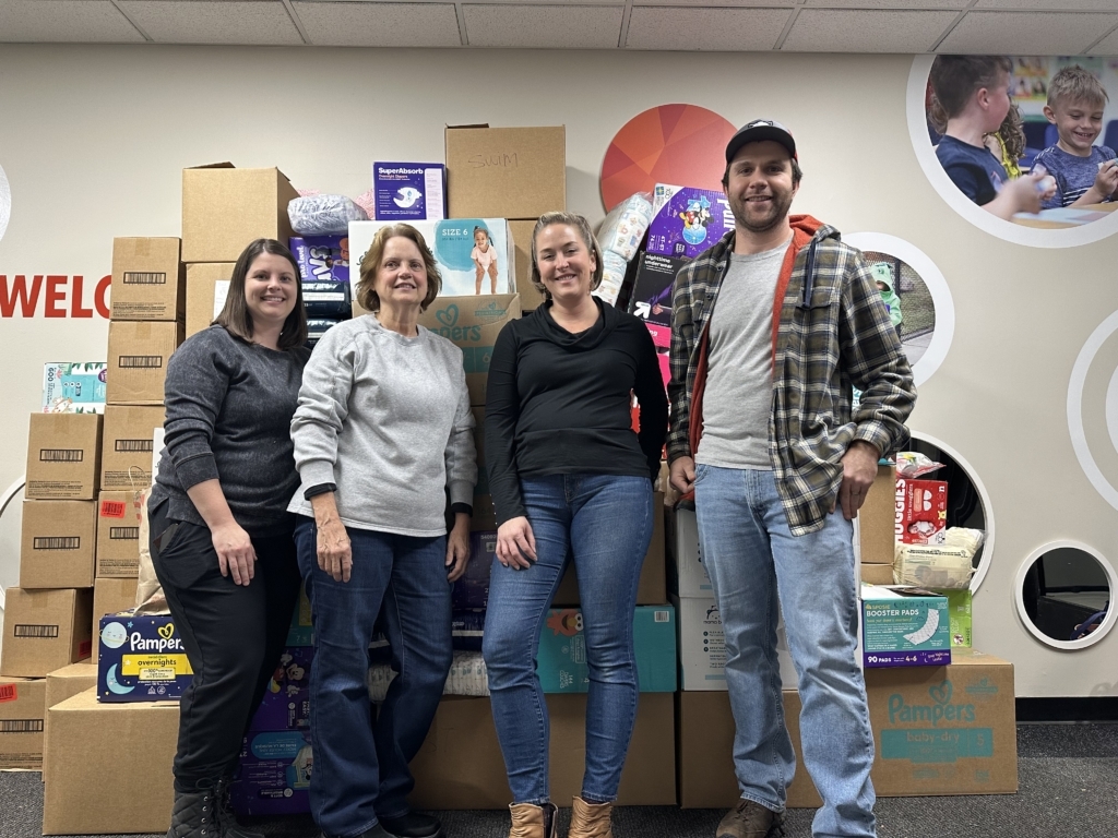 4 people stand in front of a stack of a large stack of boxes of diapers against a wall.