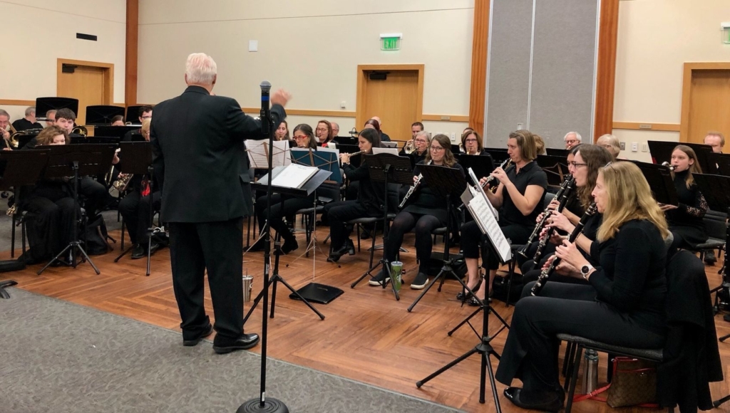Eden Prairie Community Band members, dressed in black, seated playing instruments at the arboretum while a conductor stands in the foreground with his back to the camera.