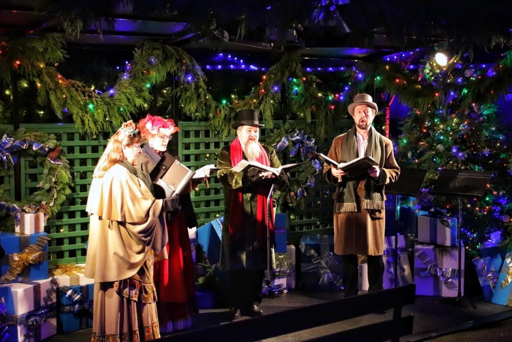 Two men and two women dressed in Victorian costumes sing Christmas carols surrounded by pine garland