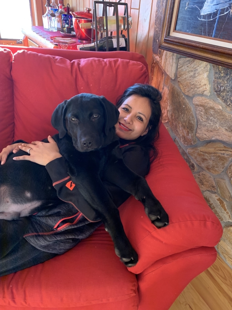 Young woman cuddles on red couch with black Lab dog.