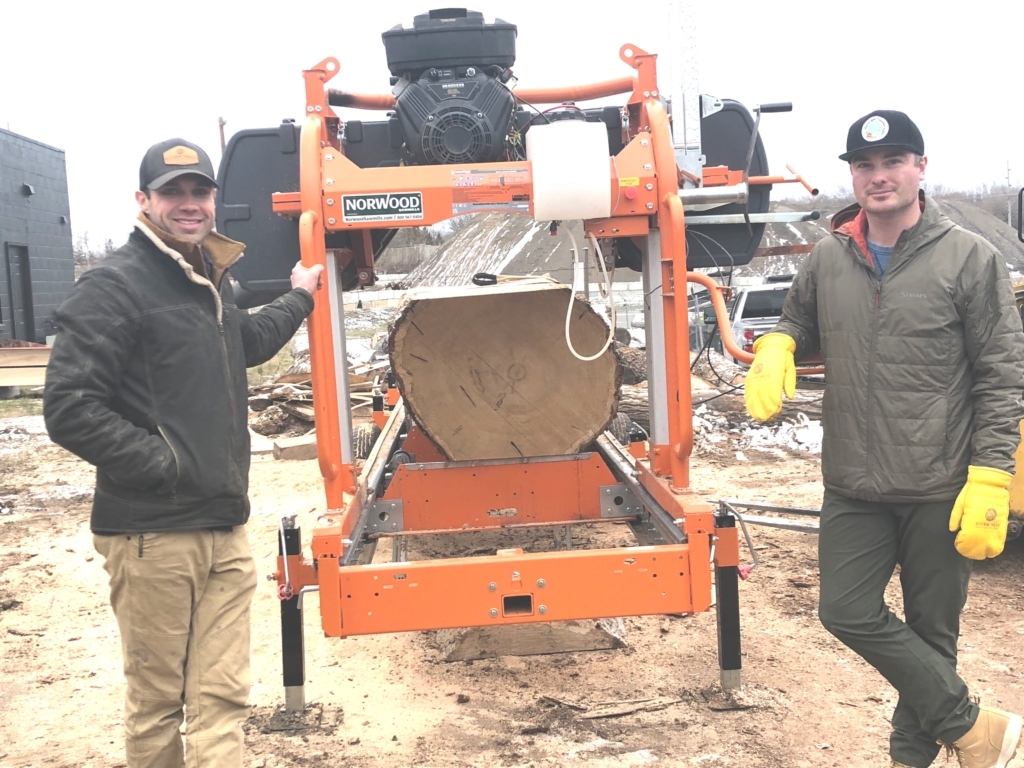 2 men posing outdoors next to orange Norwood sawmill with log in it