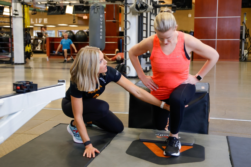 woman in red tank top does stretching exercises in gym setting while female trainer positions her leg correctly