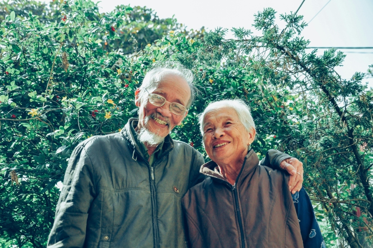Smiling elderly man and woman wearing jackets outside. He has his arm around her shoulders.