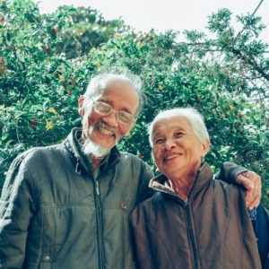 Smiling elderly man and woman wearing jackets outside. He has his arm around her shoulders.