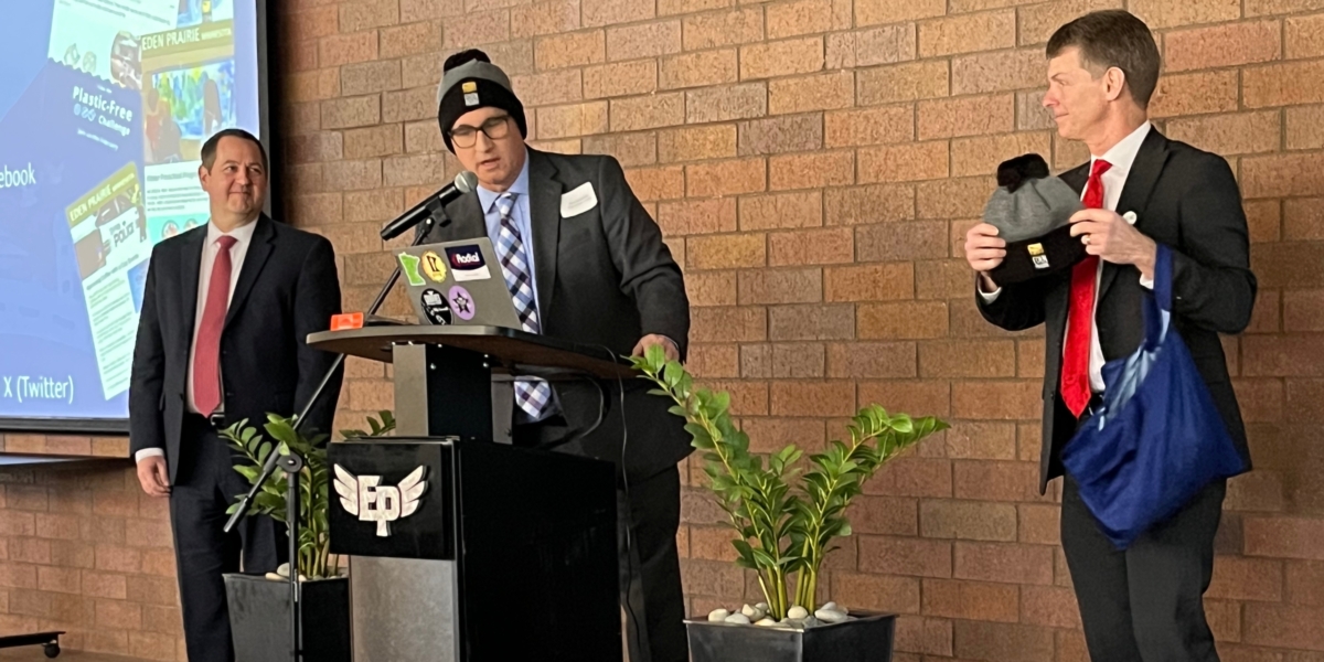 man wearing a suit and stocking cap stands at podium, another man in suit holds up a stocking cap while a third man in a suit looks on