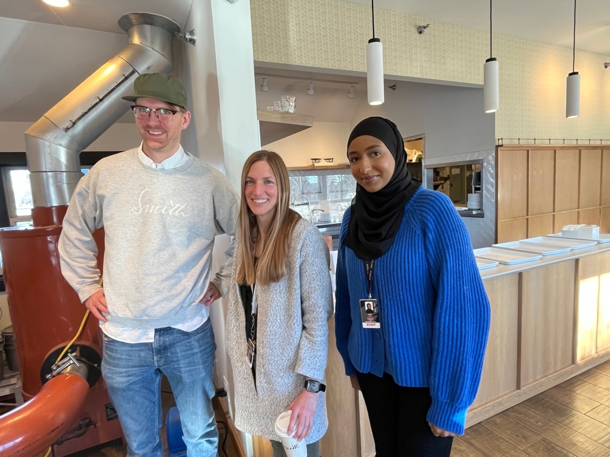 man in white sweater and ball cap, woman in white sweater, woman in blue sweater stand in front of large coffee machine in coffee shop interior