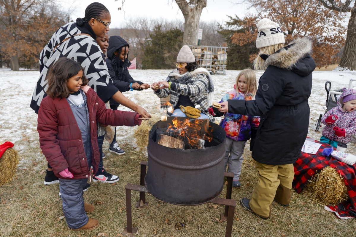 Several people toast s'mores over an outdoor fire pit in a winter landscape.