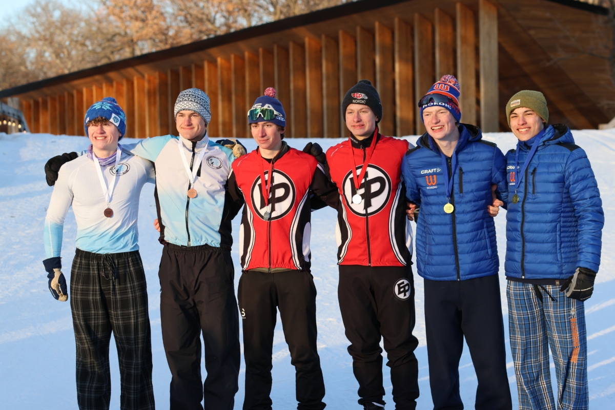 6 young men stand with arms around each other's shoulders in snowy landscape; 2 in middle wear Eden Prairie Nordic ski uniforms
