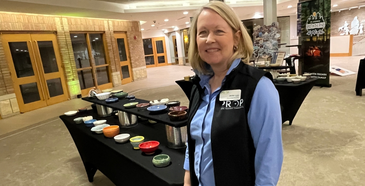 woman in blue shirt with black vest stating PROP stands in front of table of pottery bowls