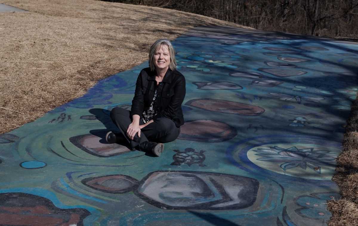 This image has an empty alt attribute; its file name is EPLN_ArtInstallations-9820-683x1024.jpg Lori Brink, Eden Prairie’s recreation services manager, stands at the River Rapids mural at Staring Lake Park. The mural, painted by artists Liv Novotny and Ramon Madden, depicts the Mississippi River and its ecosystems, ultimately leading to a slide on the playground. Photo by Jeremy Peyer