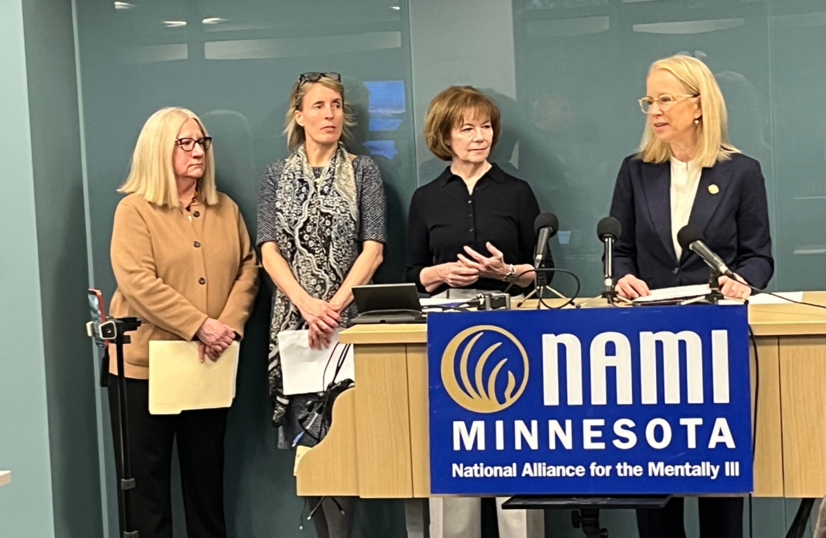 blond woman speaks at podium behind NAMI Minnesota sign while 3 other women look on