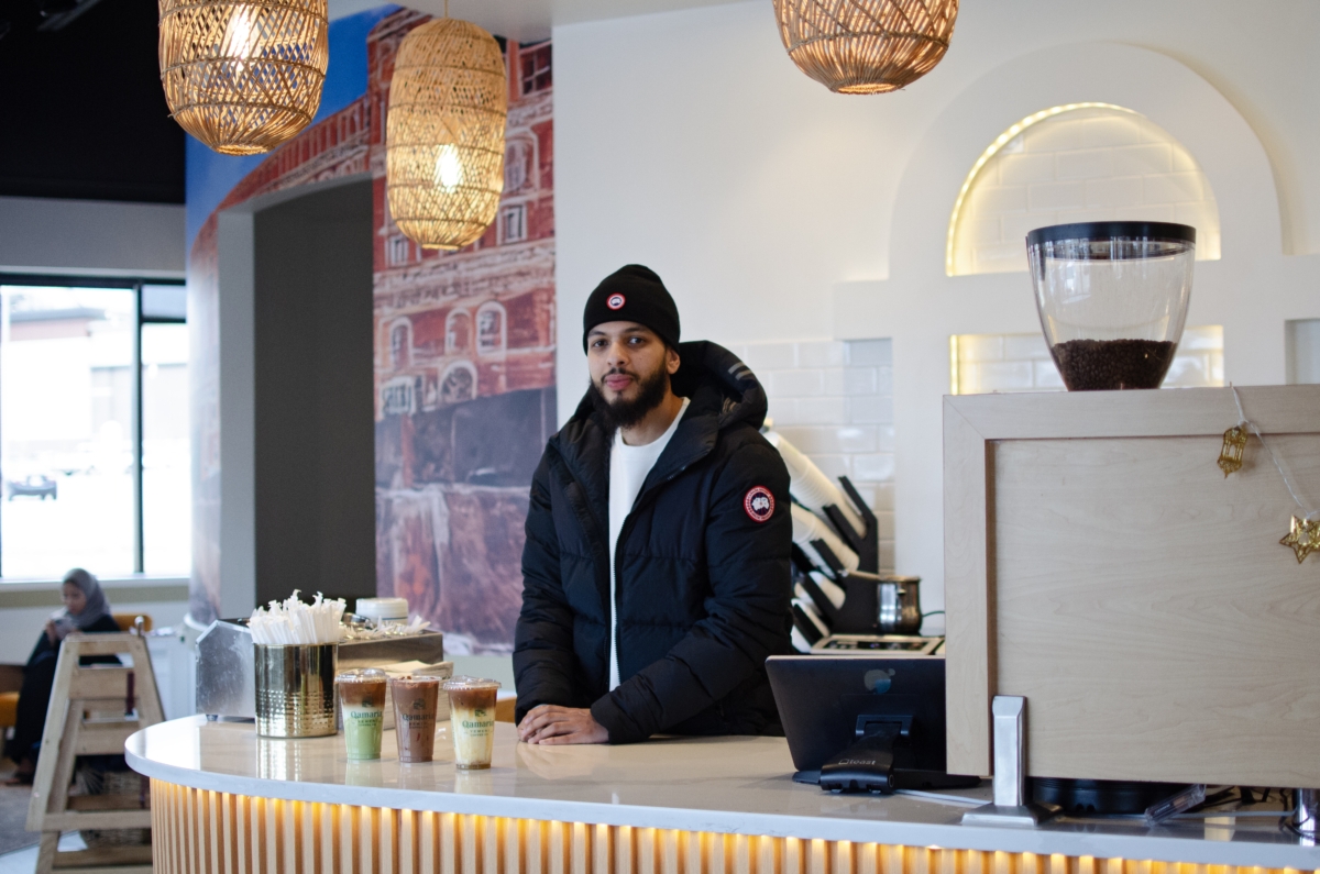Qamaria Coffee managing partner and co-owner Mohamed Hagi stands at the counter of the Eden Prairie coffee shop. surrounded by warm lighting and bright white walls. Three of their bestselling items, including two lattes, sit in front of him.