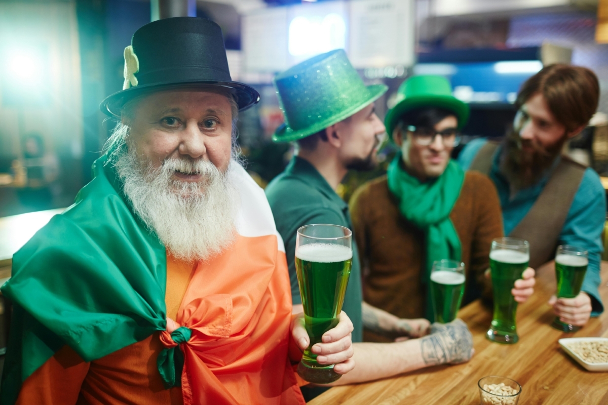 A photo of an elderly man with a beard dressed in St Patrick's Day regalia, drinking a beer