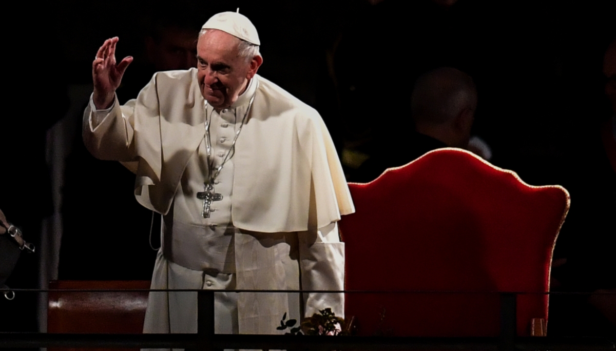 Pope Francis standing in front of red chair with hand raised in blessing