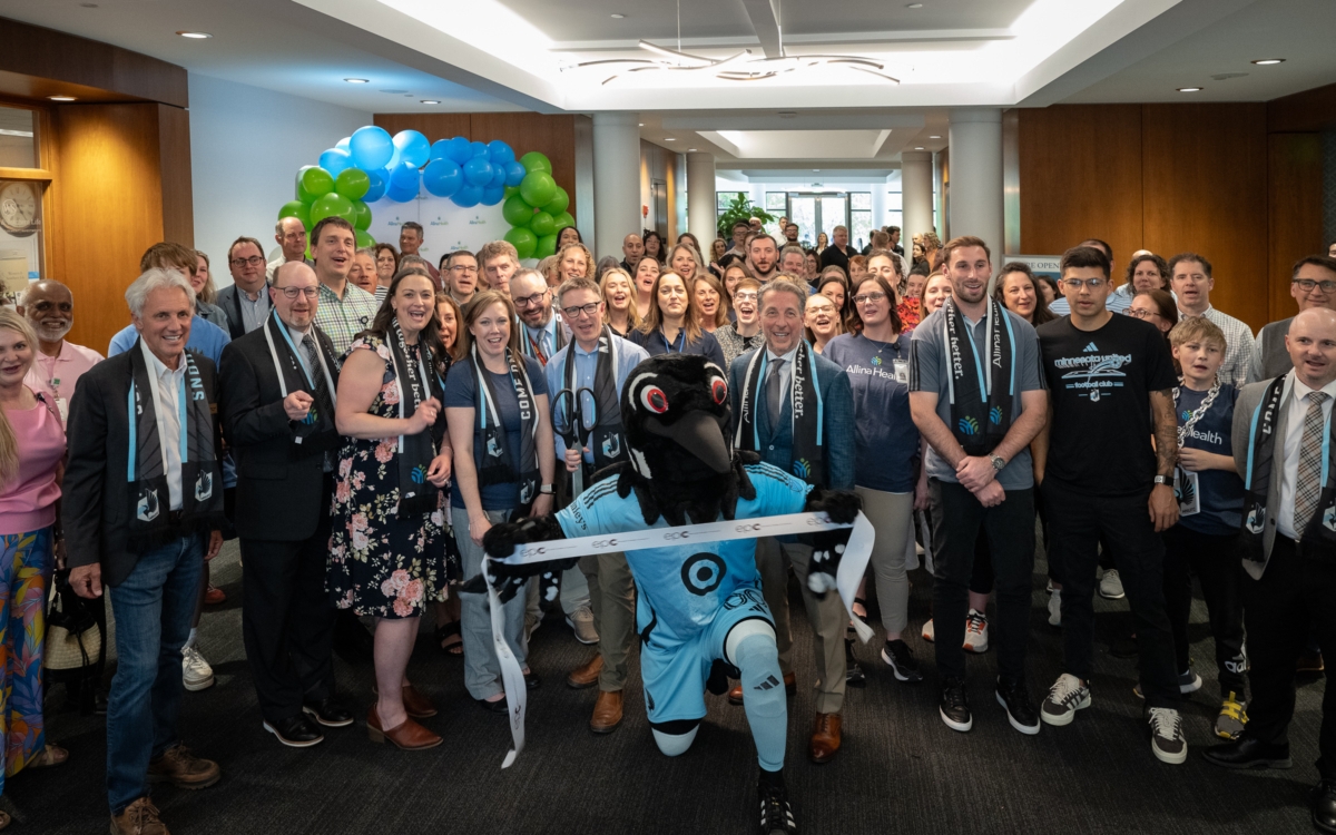 A loon mascot kneels holding a ribbon in front of a crowd of people