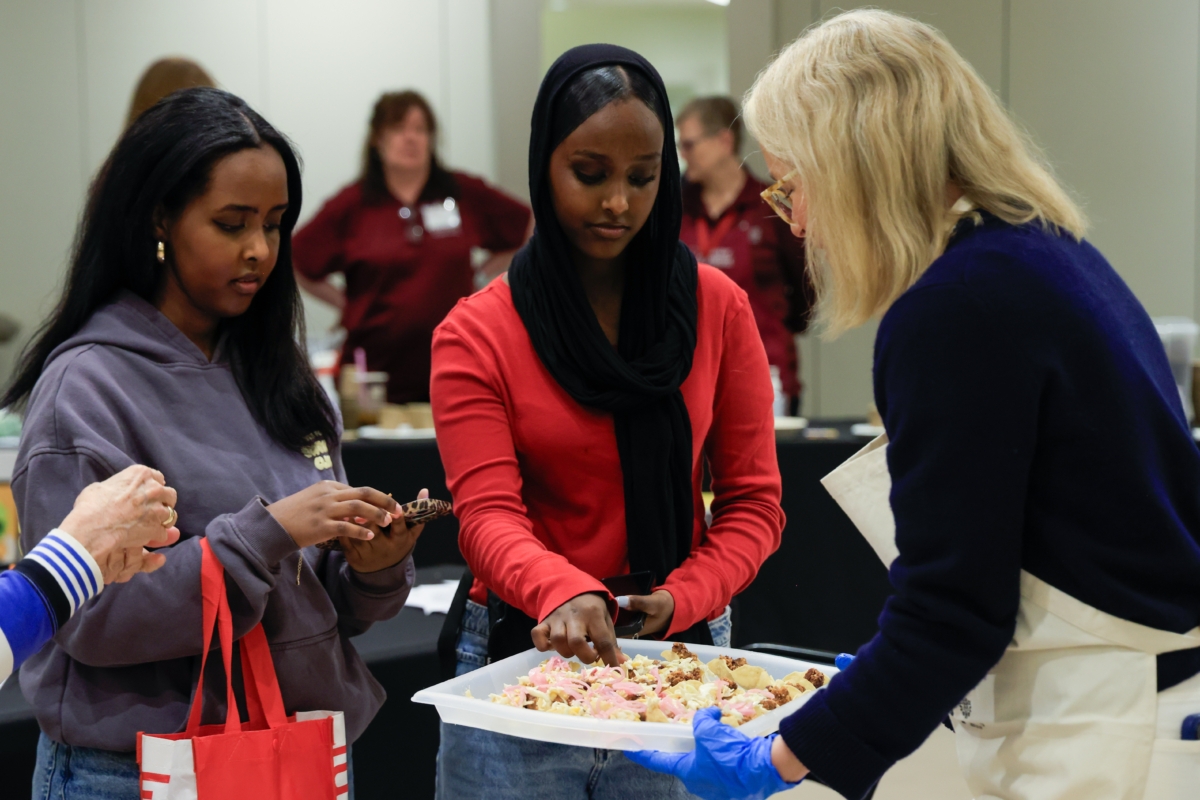 2 girls try food samples from plate held by blond woman in apron.