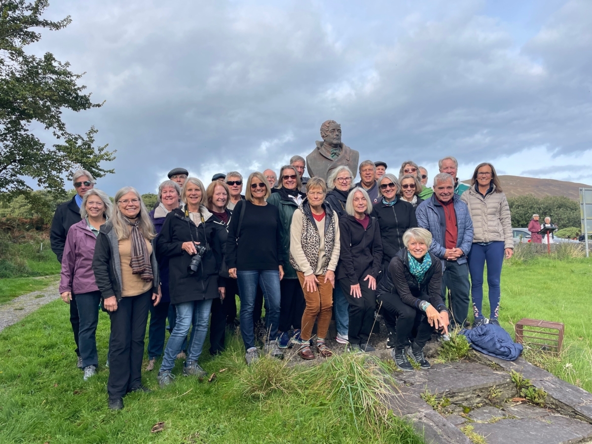 A group of 26 travelers stand on a field of green grass with a statue in front of a cloudy sky. Their trip to Ireland is one of four trips the teachers have led so far.