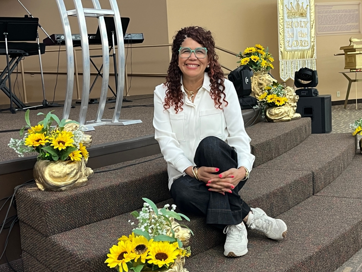 Pastor Marisol McKay, in white shirt and black pants, sits among yellow flowers on altar steps at Casa de Victorias church in Eden Prairie