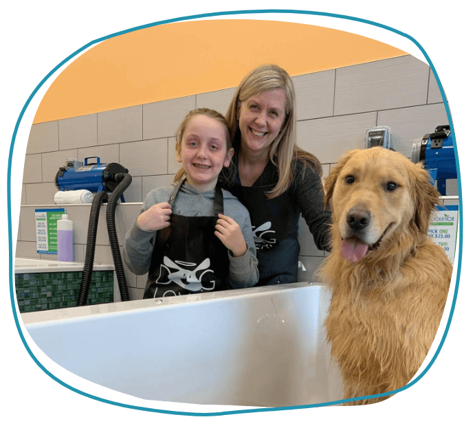 woman and girl in apron smile while damp golden retriever dog sits in tub