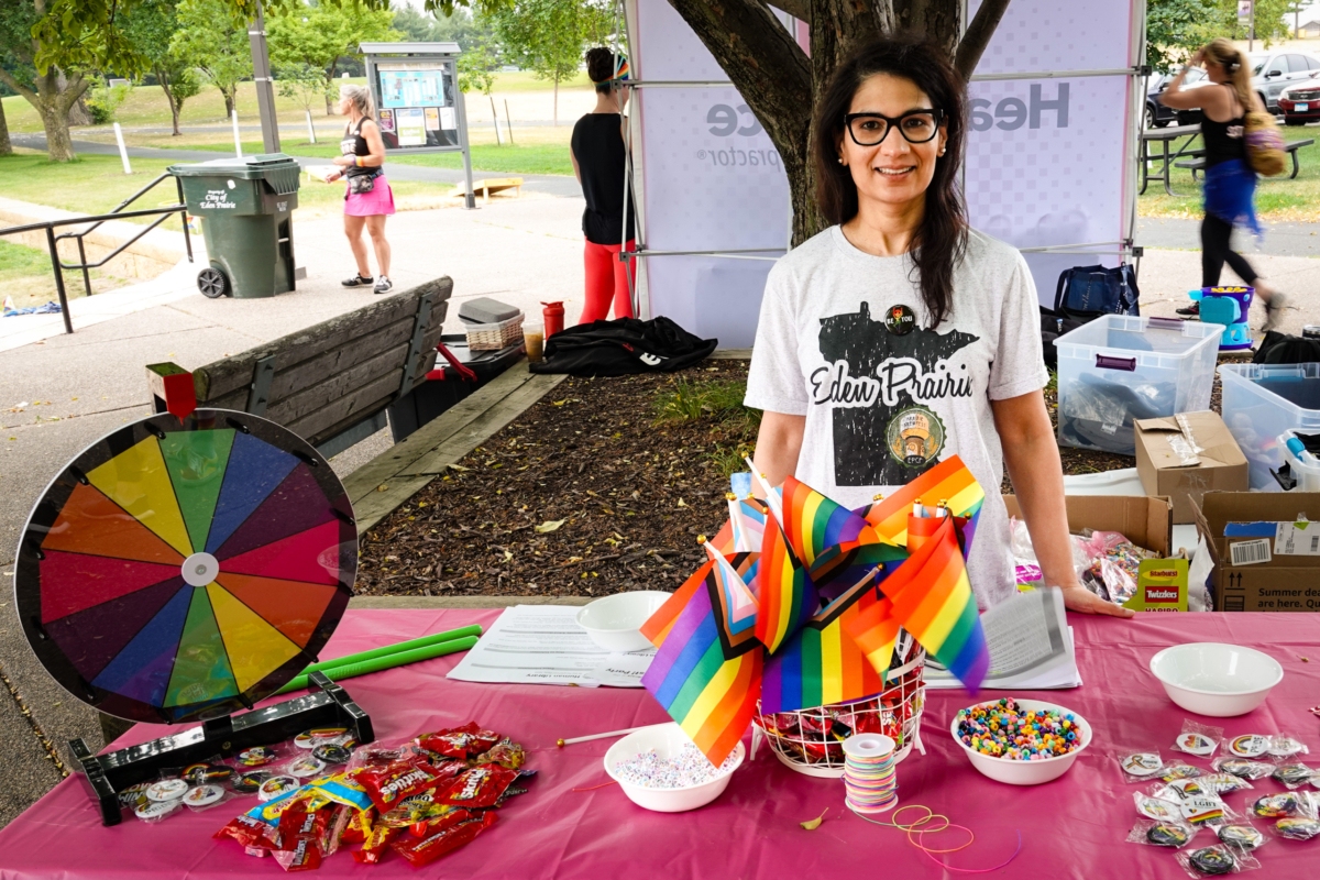 woman in Eden Prairie T-shirt stands behind table with rainbow flags at outdoor event