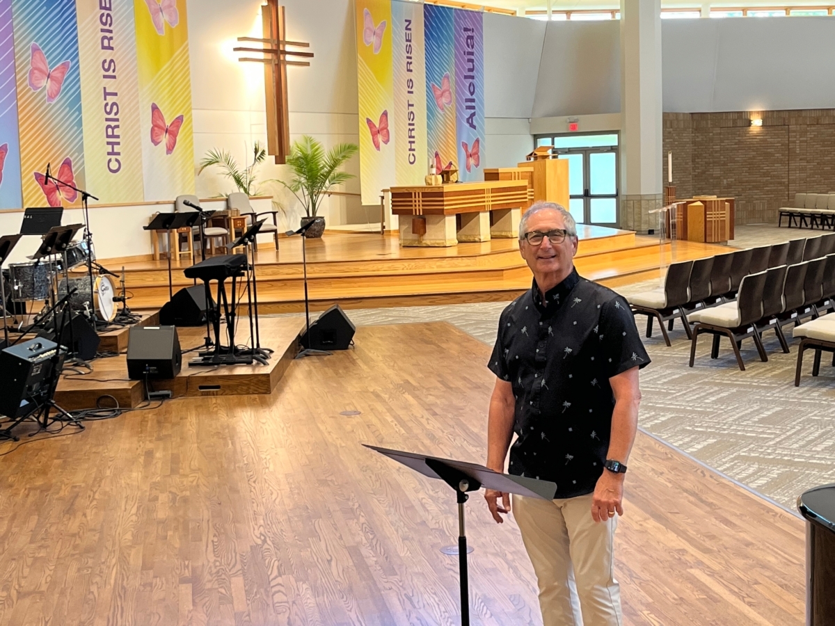 man standing at a music stand in a church setting with cross, butterfly banners on wall