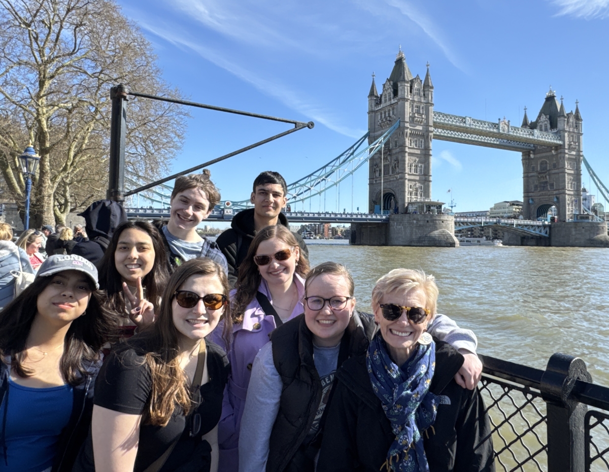 A group of seven students and their teacher stand smiling on spring break. They're in front of Tower Bridge and a blue sky.