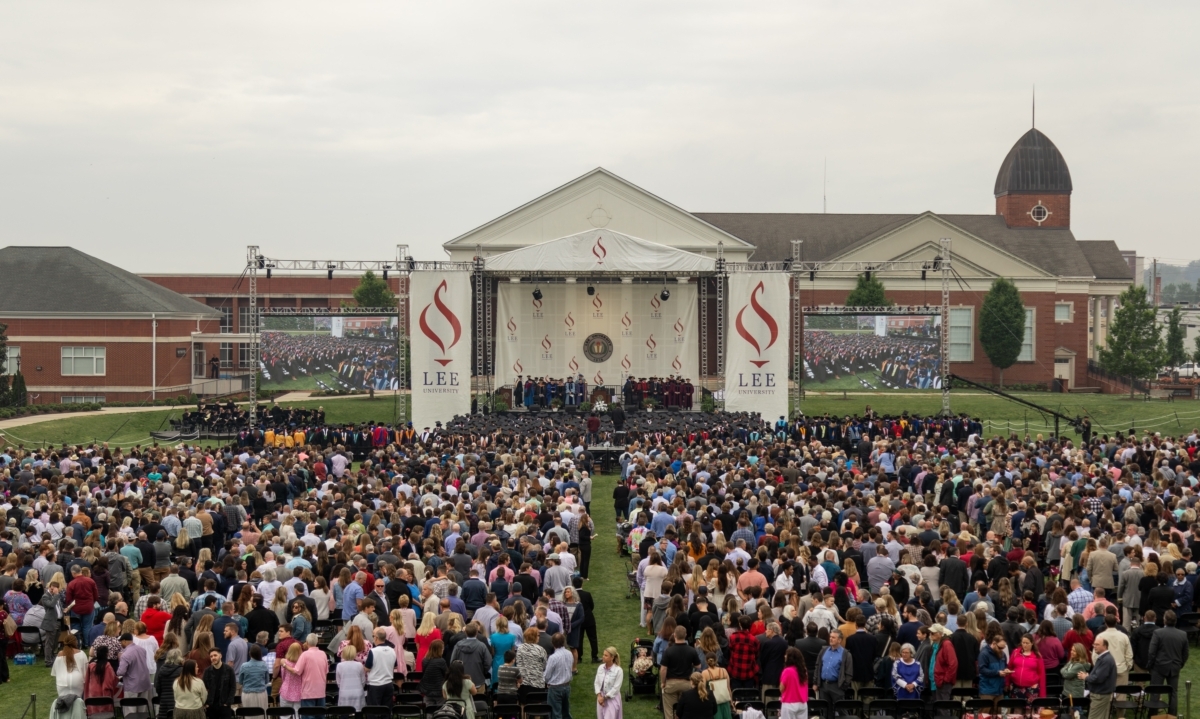 large group of people on yard of college campus as people in graduation gowns are on stage