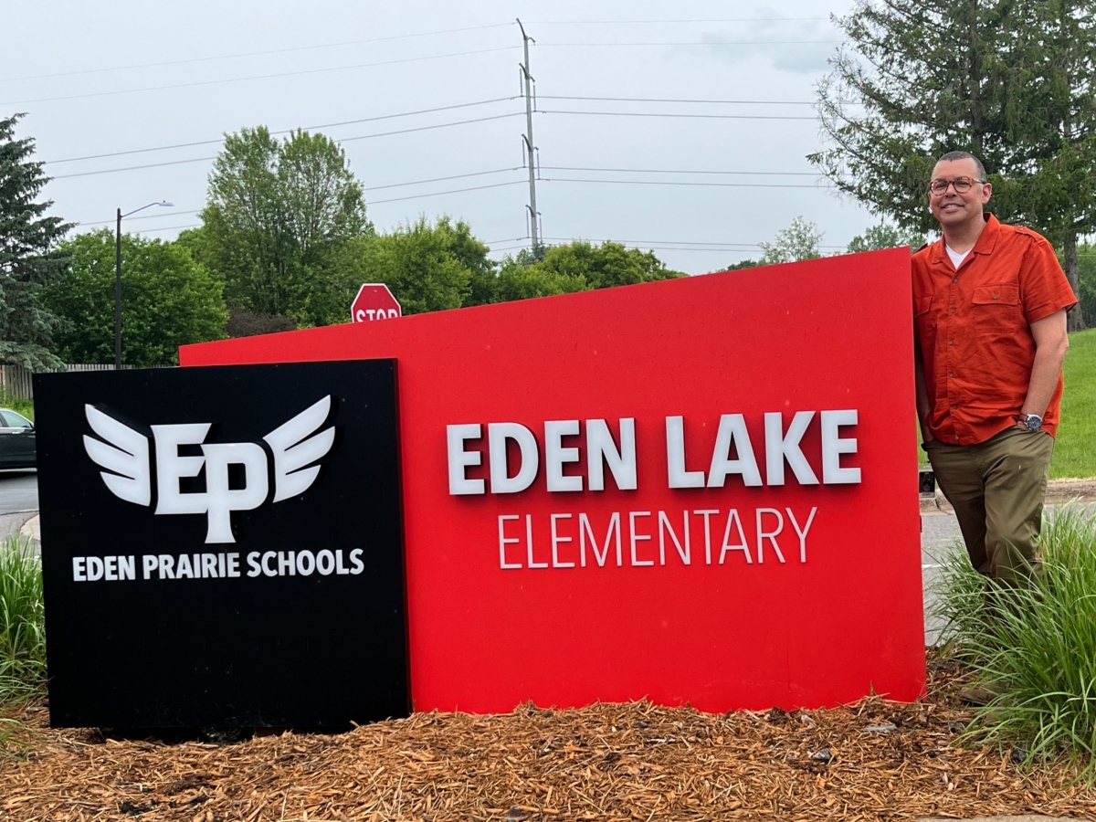 Man in red shirt and khaki pants stands next to outdoor Eden Lake Elementary sign