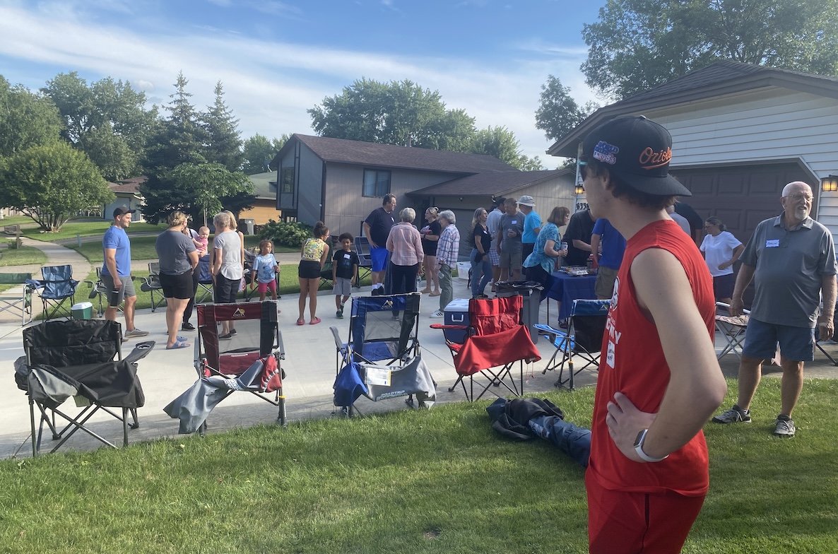 a group of people gathered near a residential driveway with lawn chairs set up