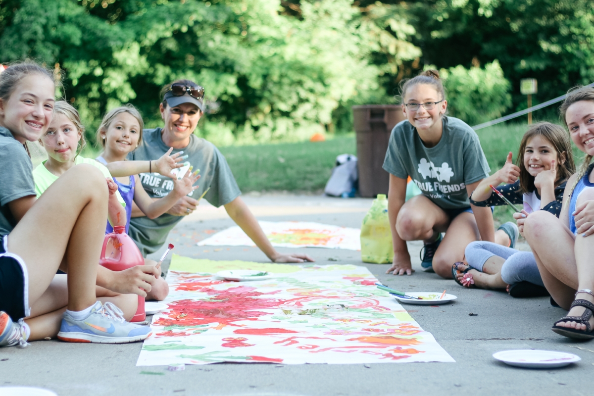 Campers and staff members sit around a large piece of paper covered in colorful paints.