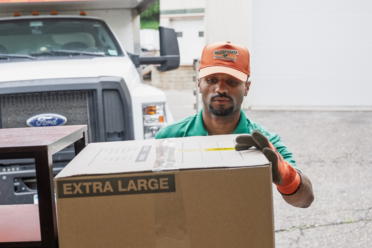 HUNKS moving employee moving large box from a moving truck to the warehouse. Wearing a baseball cap that says College HUNKS Hauling Junk & Moving.