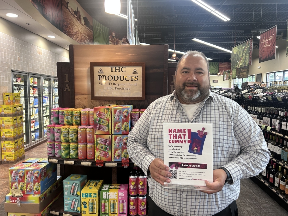 Jaime Urbina stands in an Eden Prairie Liquor store, holding a sign promoting "Name That Gummy!". On the sign, there is a picture of a purple bag with purple gummies next to it.