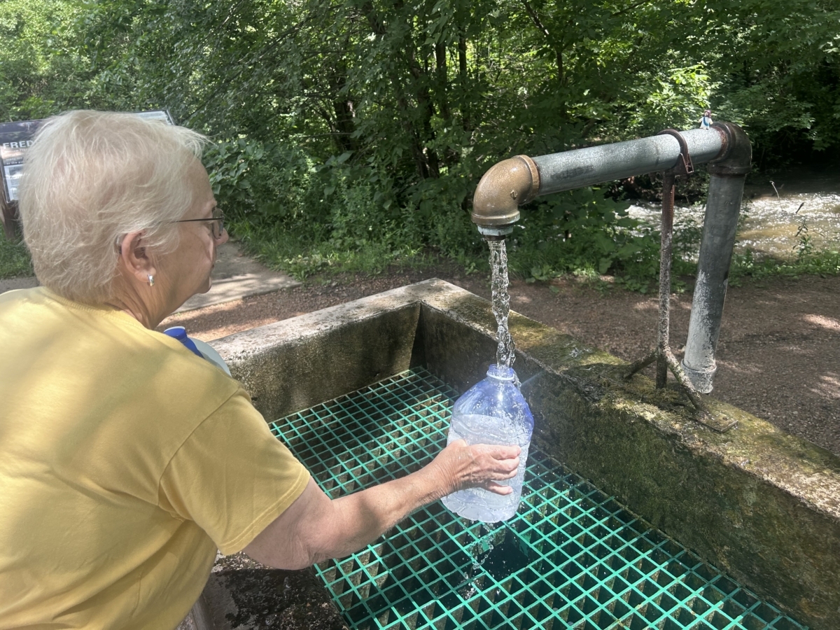 A woman holds a plastic water jug up to a metal faucet, as water fills the jug. A stone basin with a green grate is below the jug.