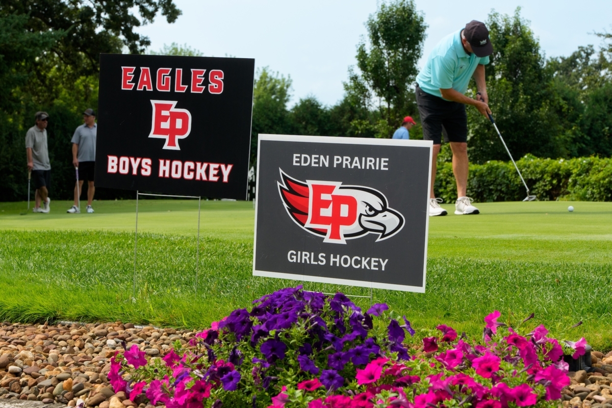 Signs for the Eden Prairie High School boys and girls hockey teams on the putting green at Bent Creek Golf Club during Monday’s joint golf tournament fundraiser, as girls head coach Steve Persian practices putting in the background.