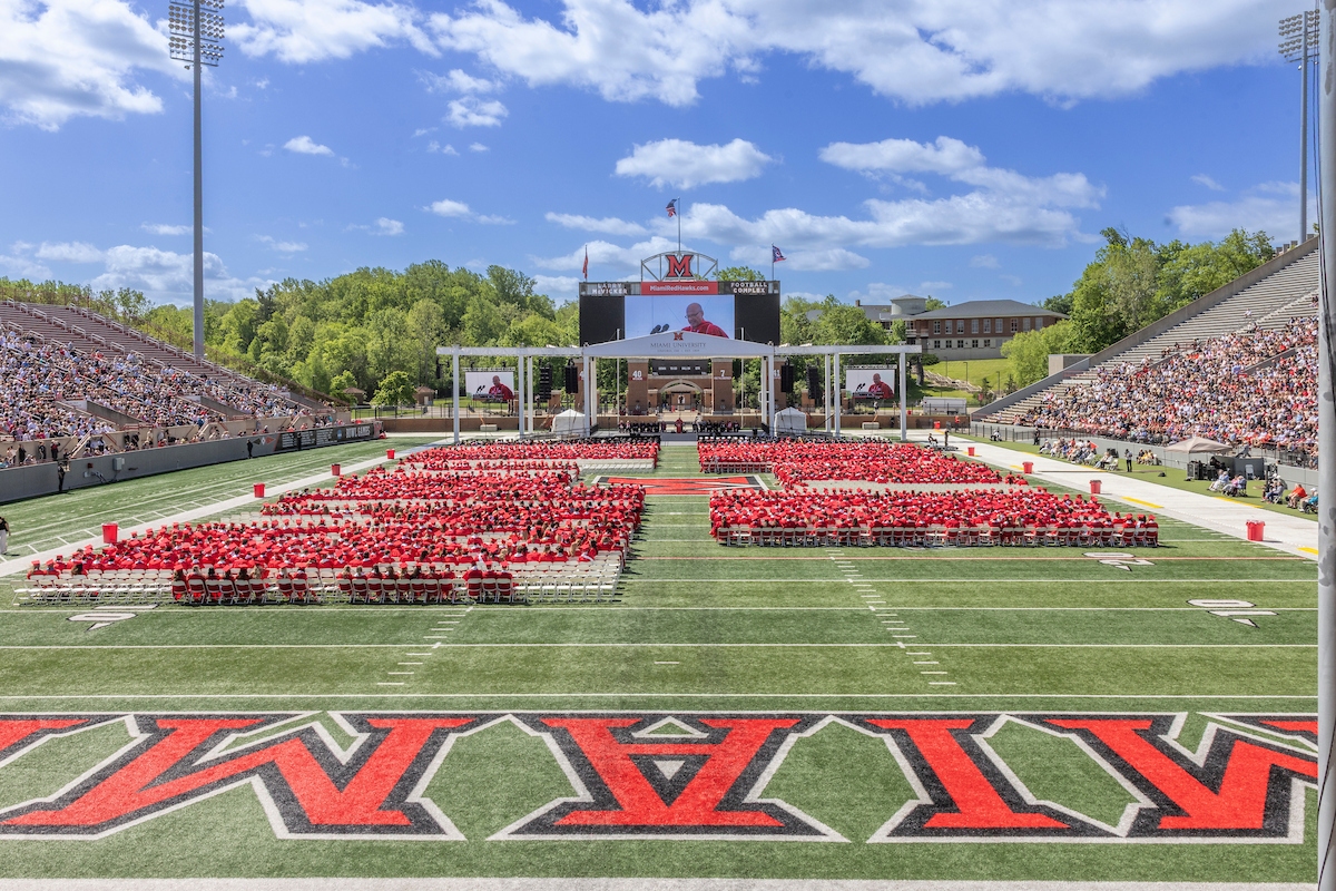 graduates in red gowns seated on an outdoor football field marked Miami