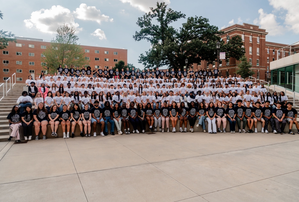 a large group of young people in black and white T-shirts seated outdoors on a college campus