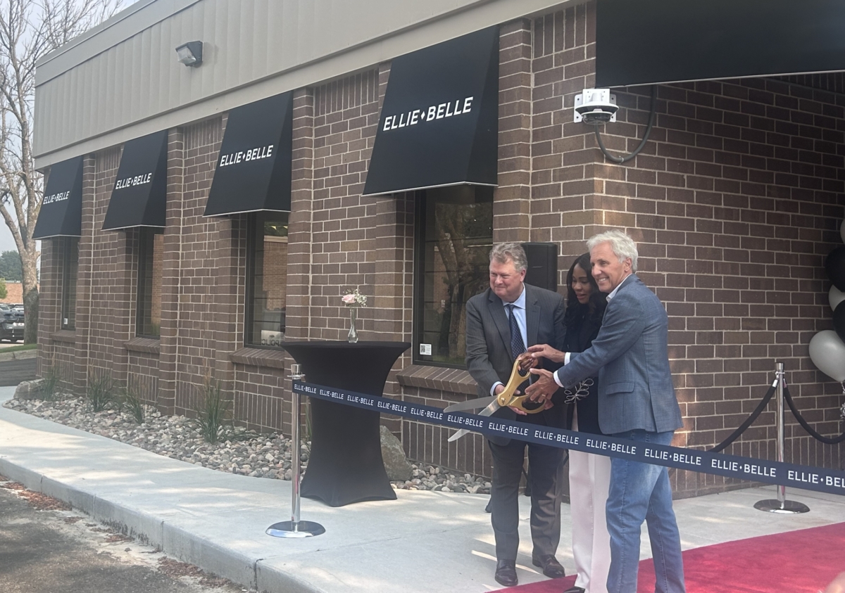 Three people stand in front of a brick building and behind a long, black ribbon. The people are standing on a red carpet and holding large gold scissors as they prepare to cut the ribbon.