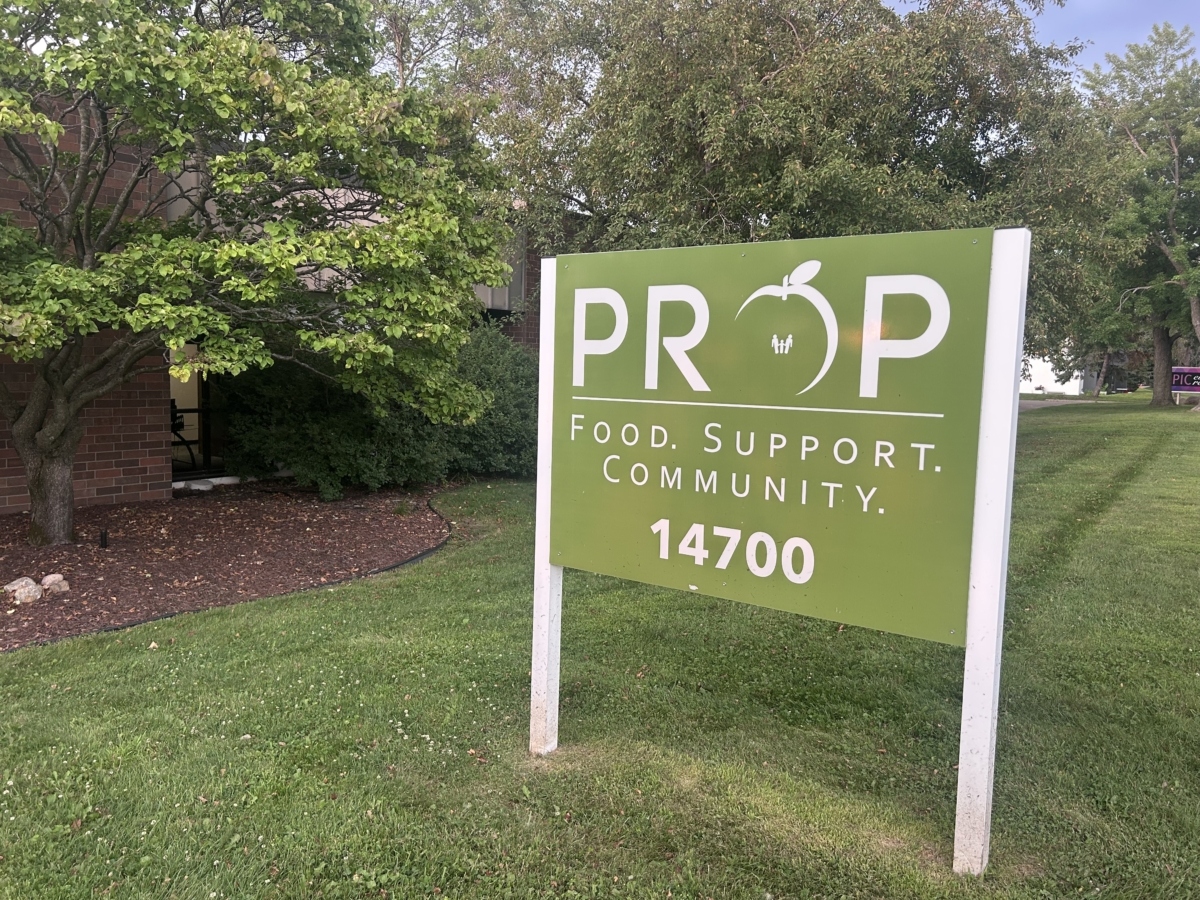 A green sign advertising PROP Food Shelf sits in front of a collection of trees.