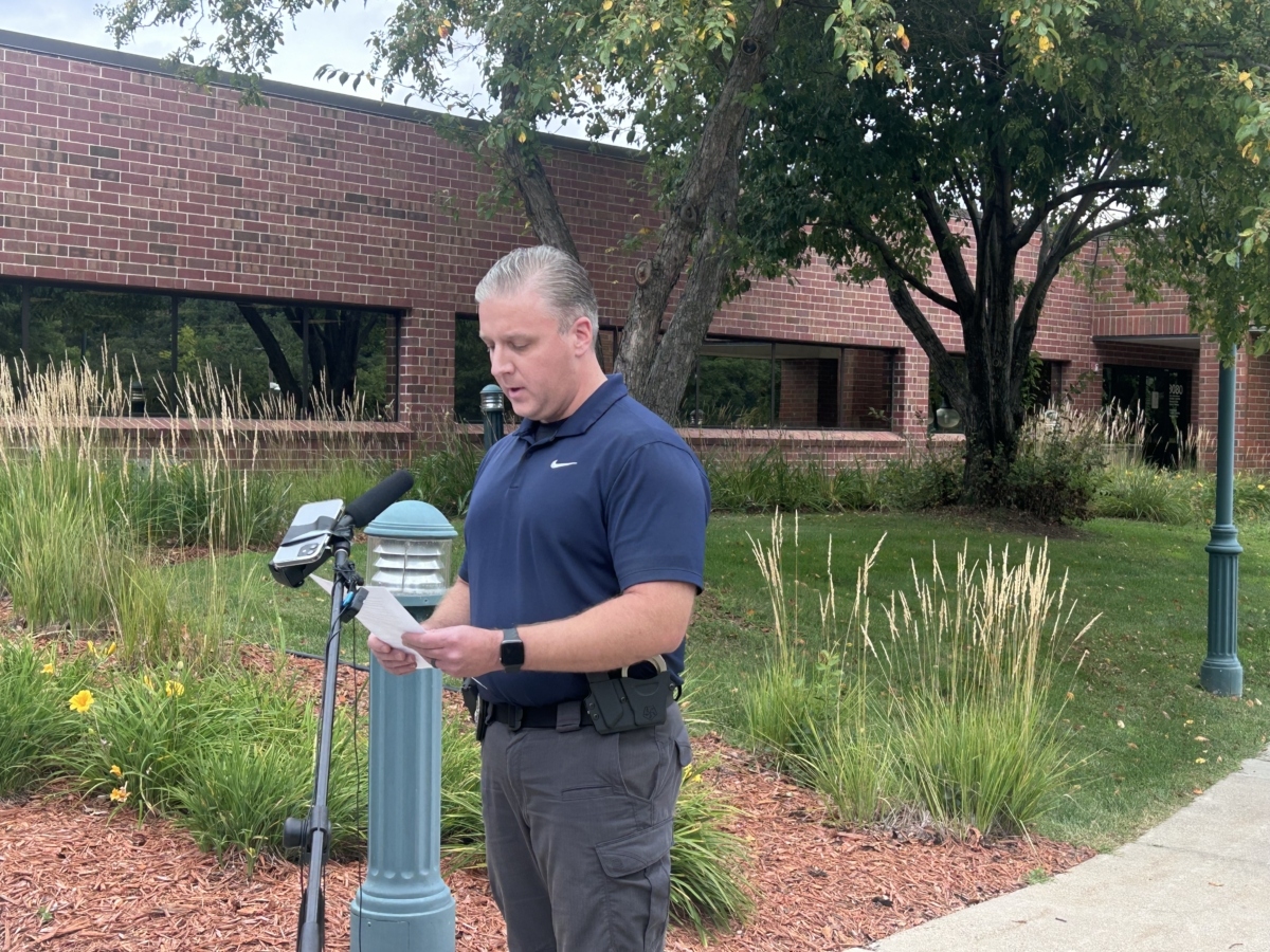 A man in a blue shirt stands in front of a microphone outside a brick building. He is reading off a sheet of paper.
