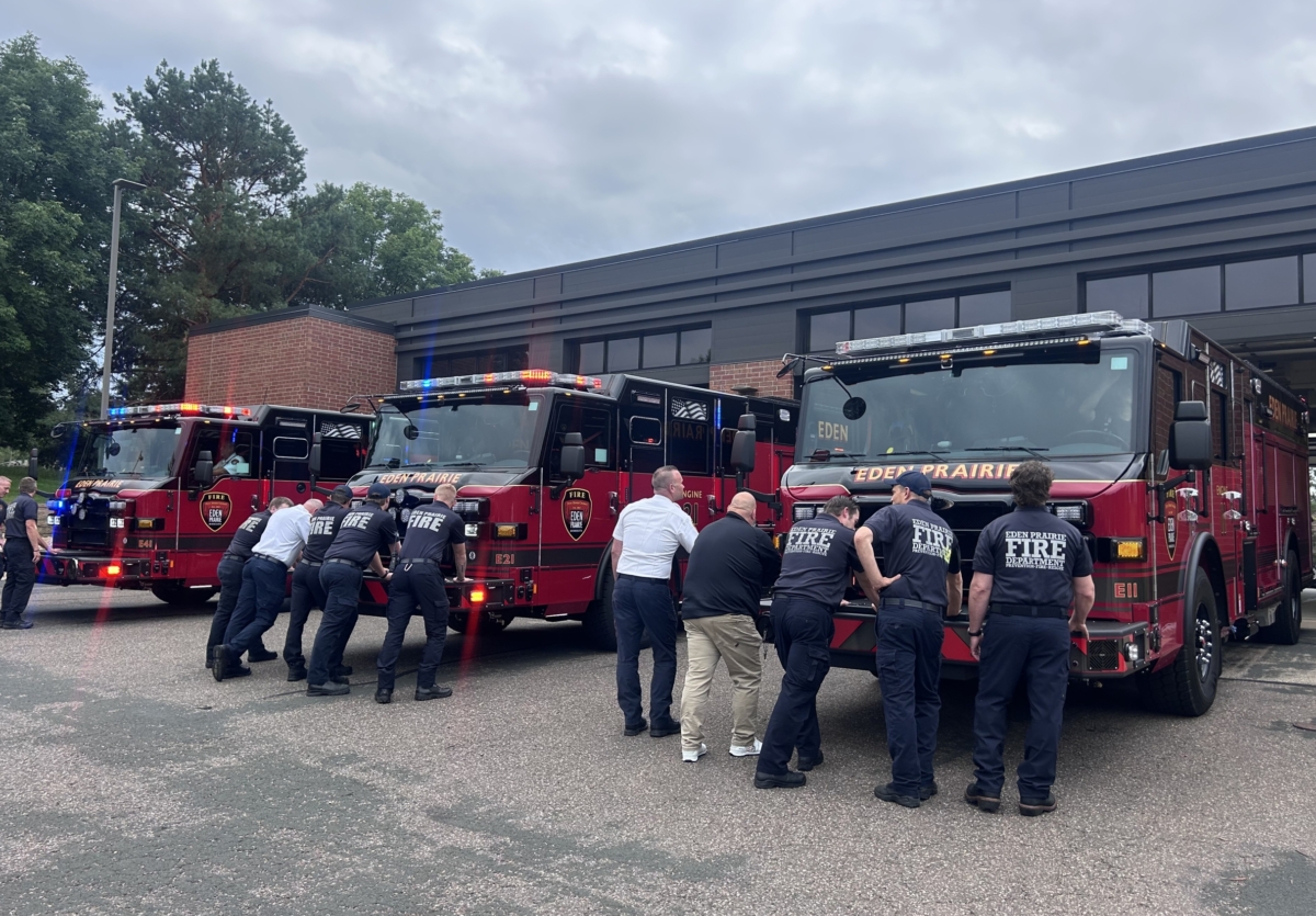 A group of firefighters pushes in three red fire trucks outside a brick building.