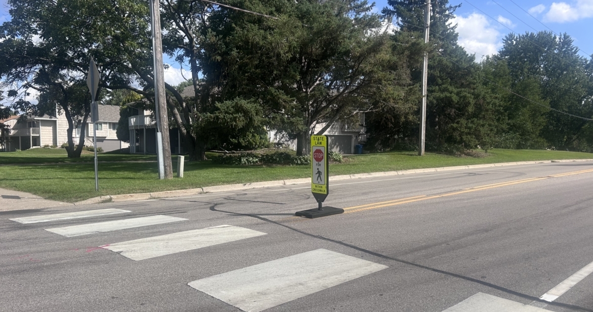 A crosswalk with a yellow sign reading "State Law Stop For Pedestrians Within Crosswalk" sits ahead of trees.