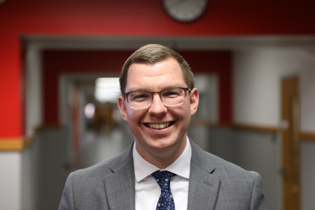 Mike Ongie smiles for a photo. He is wearing a suit and smiling in a school hallway.