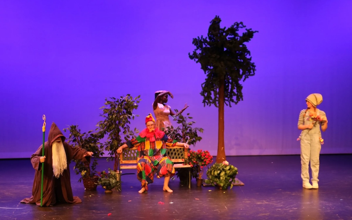 Four student actors on stage. One student is wearing a colorful, patchwork outfit and sitting on a bench, another is in a wizard outfit, one is in overalls and a hat, and the fourth is behind a bench miming an action. Fake plants and trees are onstage.