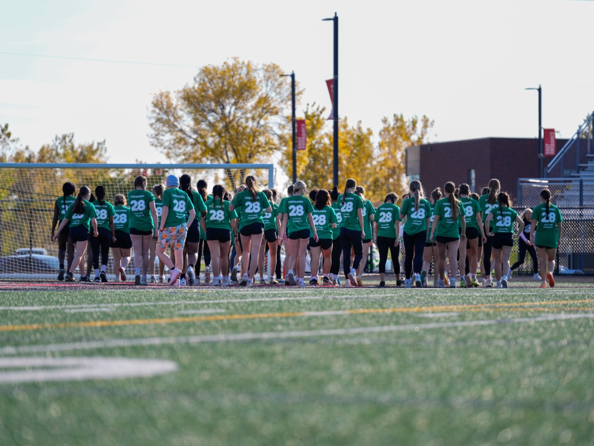 A group of freshmen participants lining up on the football field. They are all wearing matching green shirts with the number "28" and the words "Freshmen" and "Play for Possibility" written on the back.