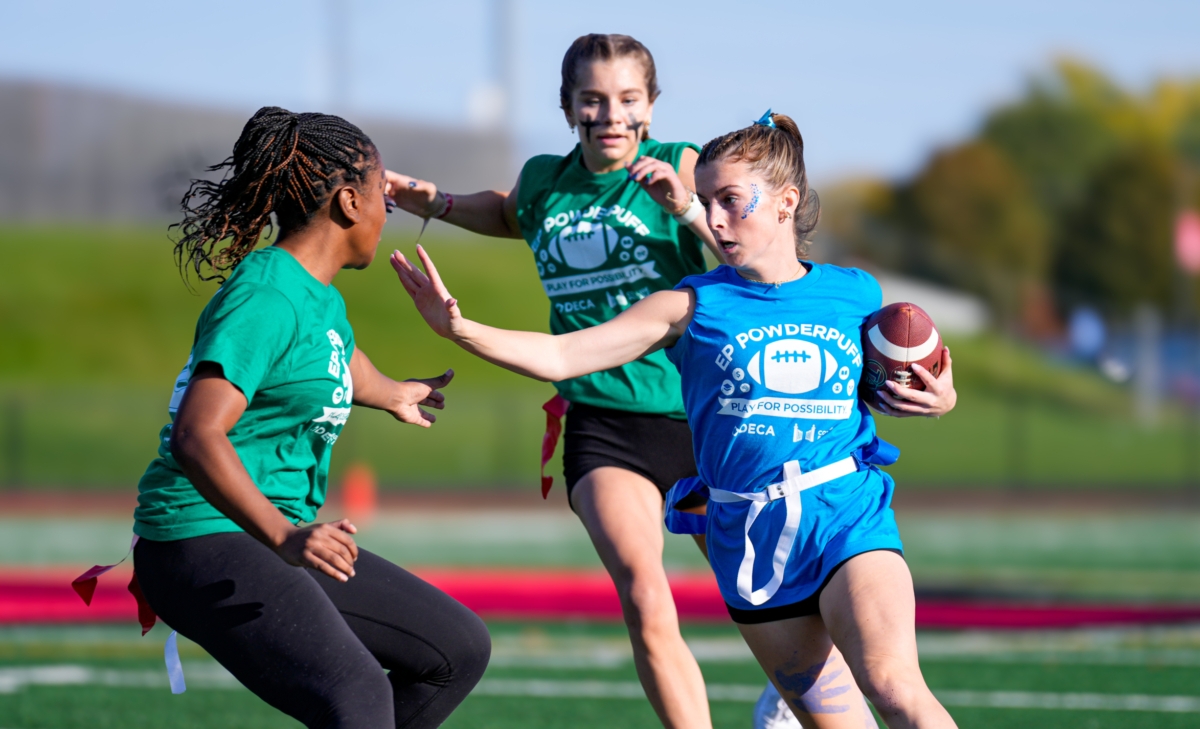 A student in a blue Powderpuff shirt runs with a football and attempts to block another student in a green Powderpuff shirt. Another student is running behind them.