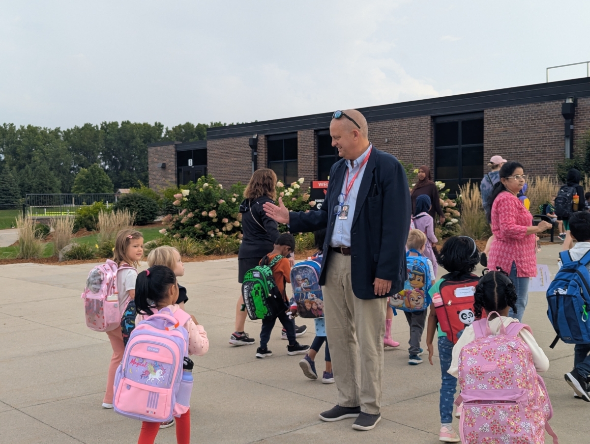 Forest Hills Associate Principal Thomas Walters smiling as he offers a high five to a student walking into Forest Hills Elementary School. There are groups of children and parents walking around them.