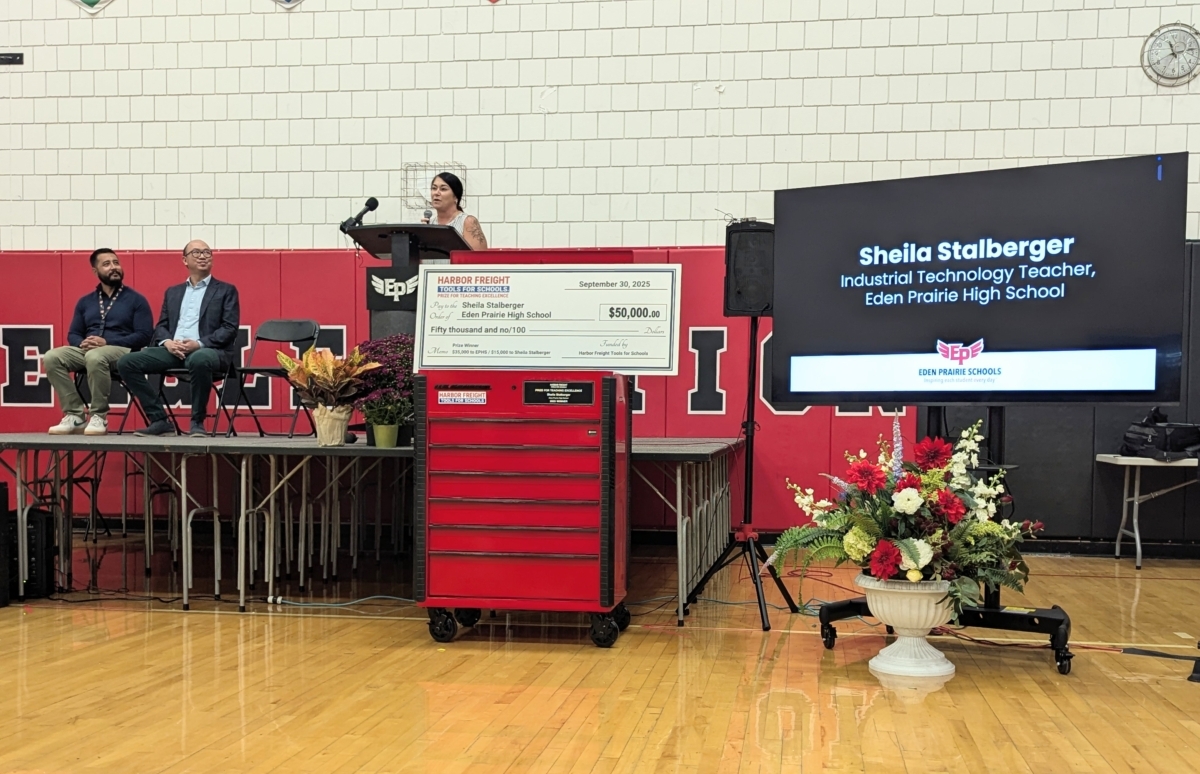 Sheila Stalberger speaks at a podium with a large check for $50,000 and a red toolkit sitting in front of her. On her left is a TV screen that says her name and her position as an industrial technology teacher at Eden Prairie High School. On her right is Tae Kang with Harbor Freight Tools for Schools and Jaysen Anderson, Eden Prairie High School's principal.